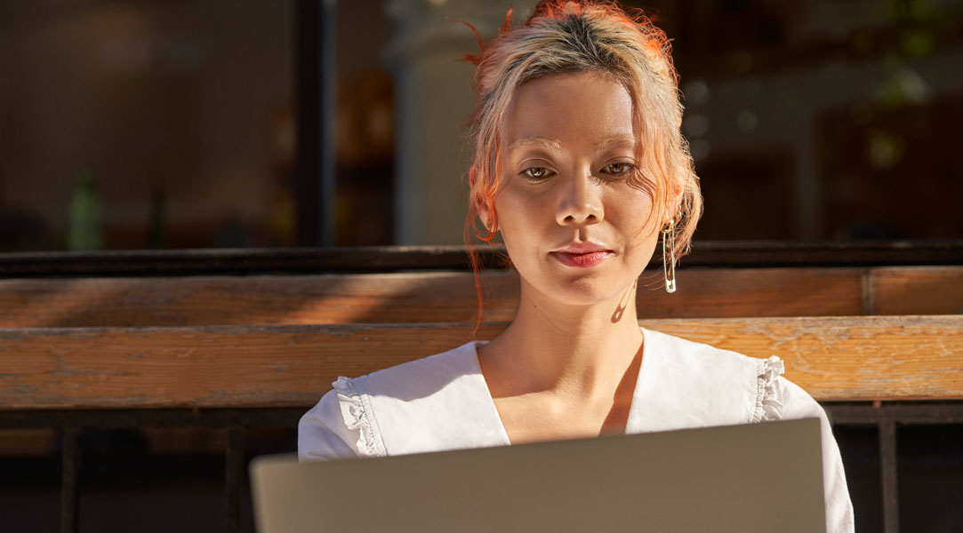 Eine Frau sitzt auf einer Bank in der Sonne und sieht auf einen Laptop.