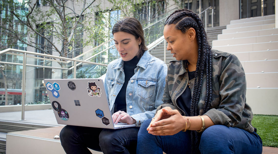 Nahaufnahme von zwei weiblichen Entwicklern, die zusammenarbeiten, während sie remote arbeiten. Eine Entwicklerin hat ihren Surface-Laptop mit Aufklebern personalisiert.