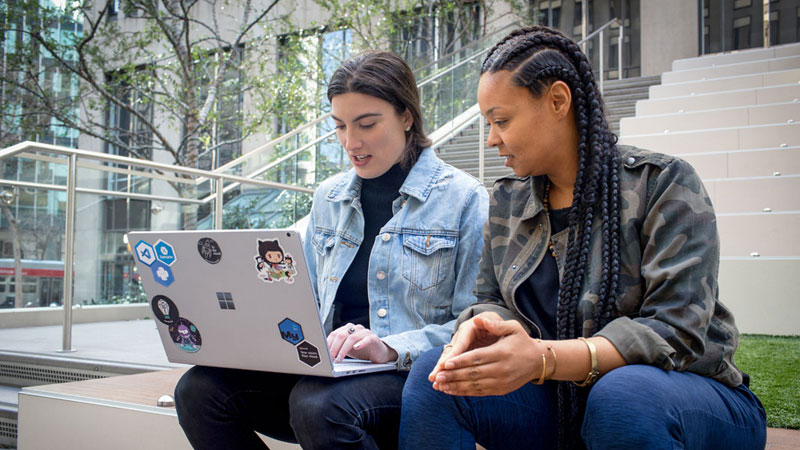 Nahaufnahme von zwei weiblichen Entwicklern, die zusammenarbeiten, während sie remote arbeiten. Eine Entwicklerin hat ihren Surface-Laptop mit Aufklebern personalisiert.